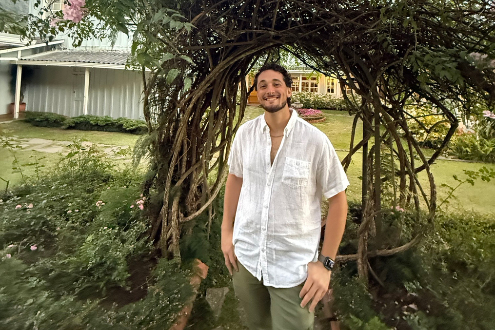 Person standing beneath a twisting flowering archway in a garden, smiling at the camera during the evening.