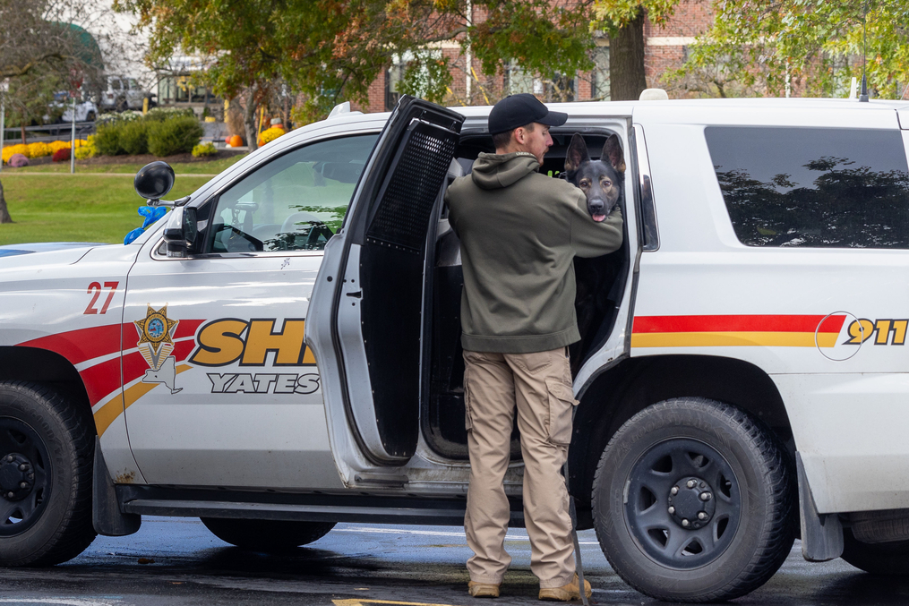 A law enforcement handler lifts a police dog from the back of a sheriff's patrol SUV parked outdoors. The dog looks toward the camera while autumn trees and a grassy area are visible in the background.